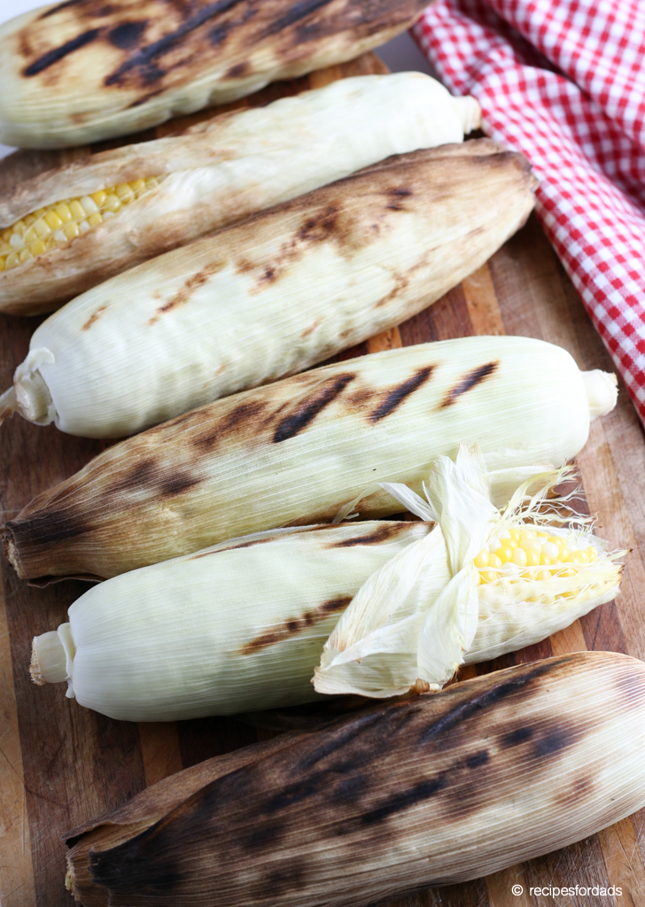 Corn on the cob with husks on, served on cutting board with a red and white checkered napkin