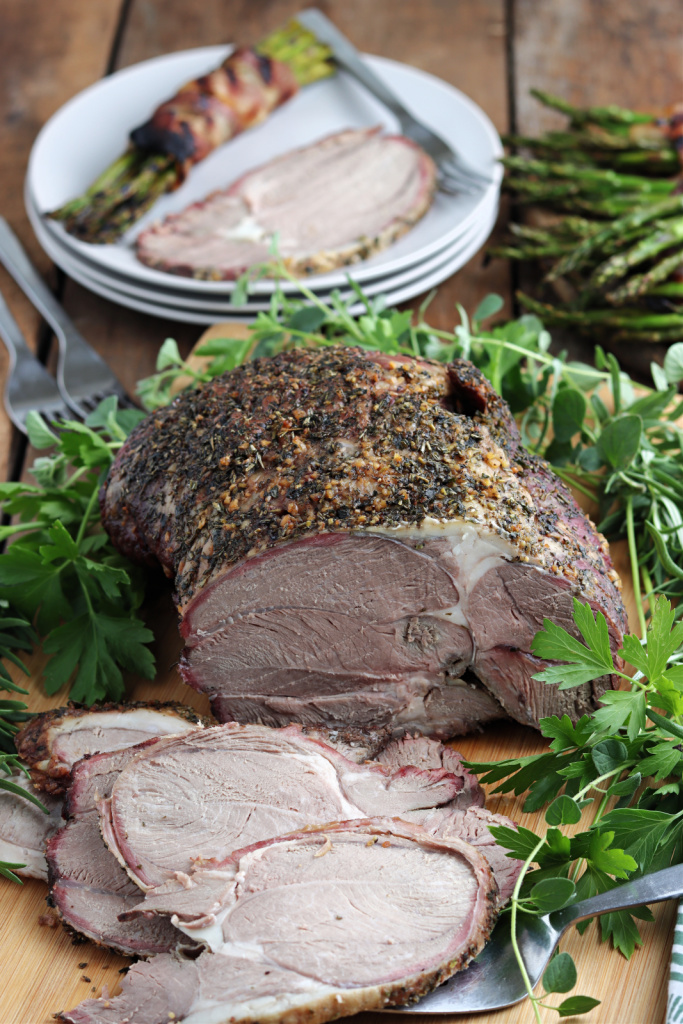 smoked leg of lamb serve on wood board with fresh herbs with asparagus wrapped in bacon