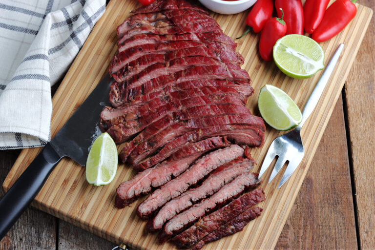smoked flank steak sliced on cutting board with knife and serving fork